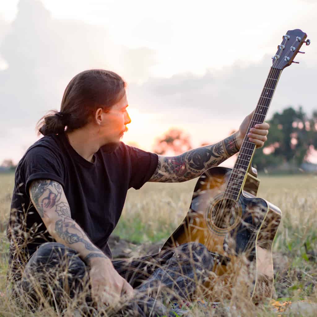 Man in a Field holding a Guitar with Bold and Colorful Sleeve Tattoos | selftattoo.com