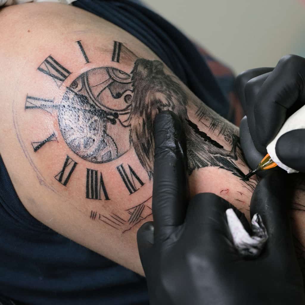 Black Linework of a Clock Face Tattoo with Roman Numerals and Visible Internal Gears Next to a Raven | selftattoo.com