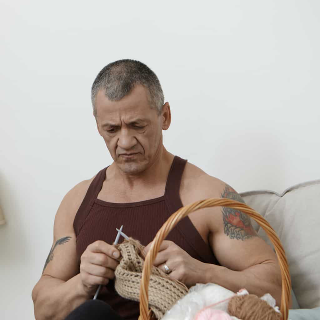 Tough and Muscular Man with Tattoos engaged in Knitting portraying a Humorous Personality | selftattoo.com